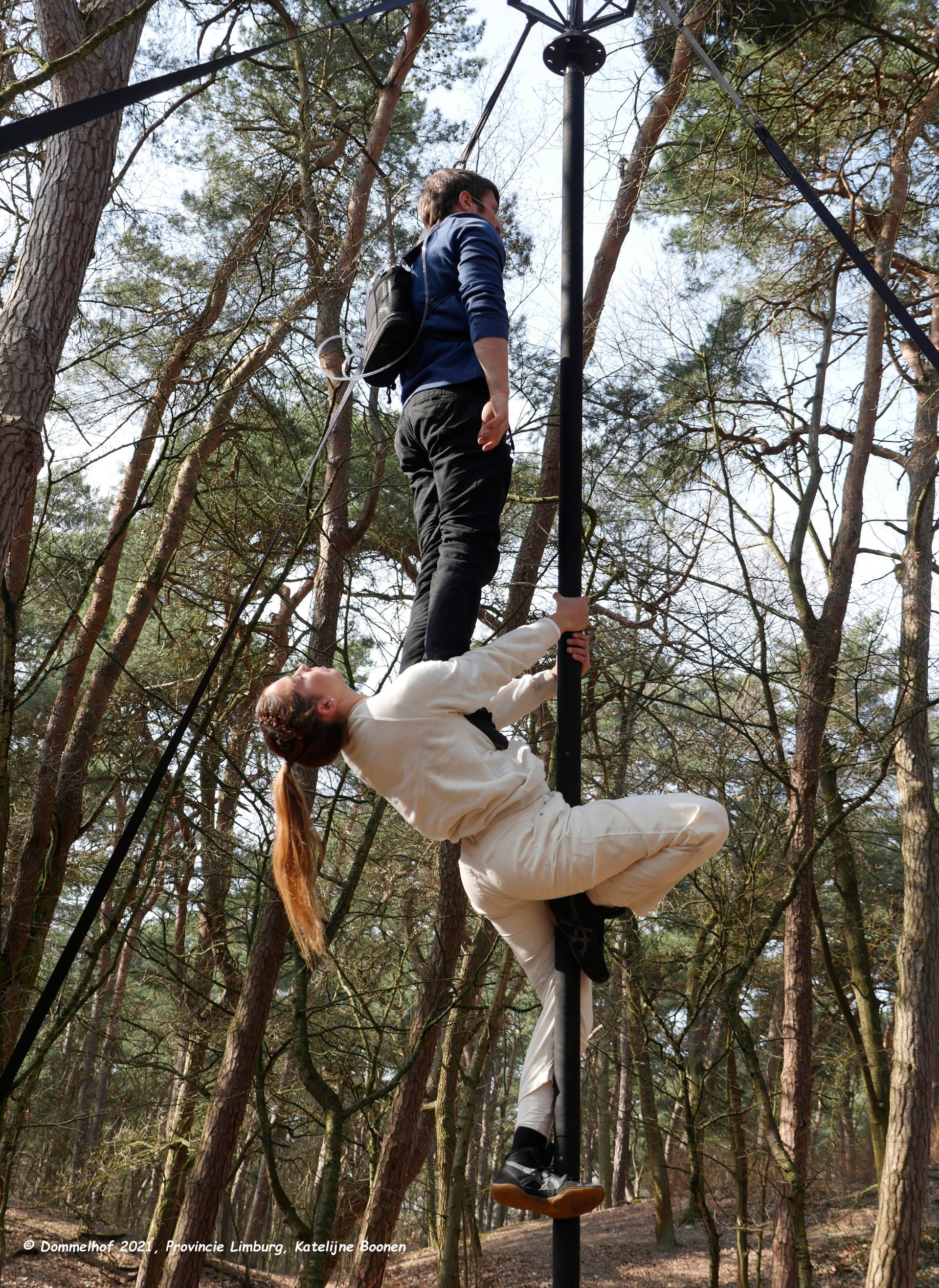 In einem Wald ist ein Chinesischer Mast aufgebaut. Daran zwei Artist*innen: Eine Person mit Sauerstofflasche auf dem Rücken steht auf der Brust der Akrobatin, die sich unter ihr mit Händen und Füßen an dem Mast festhält.