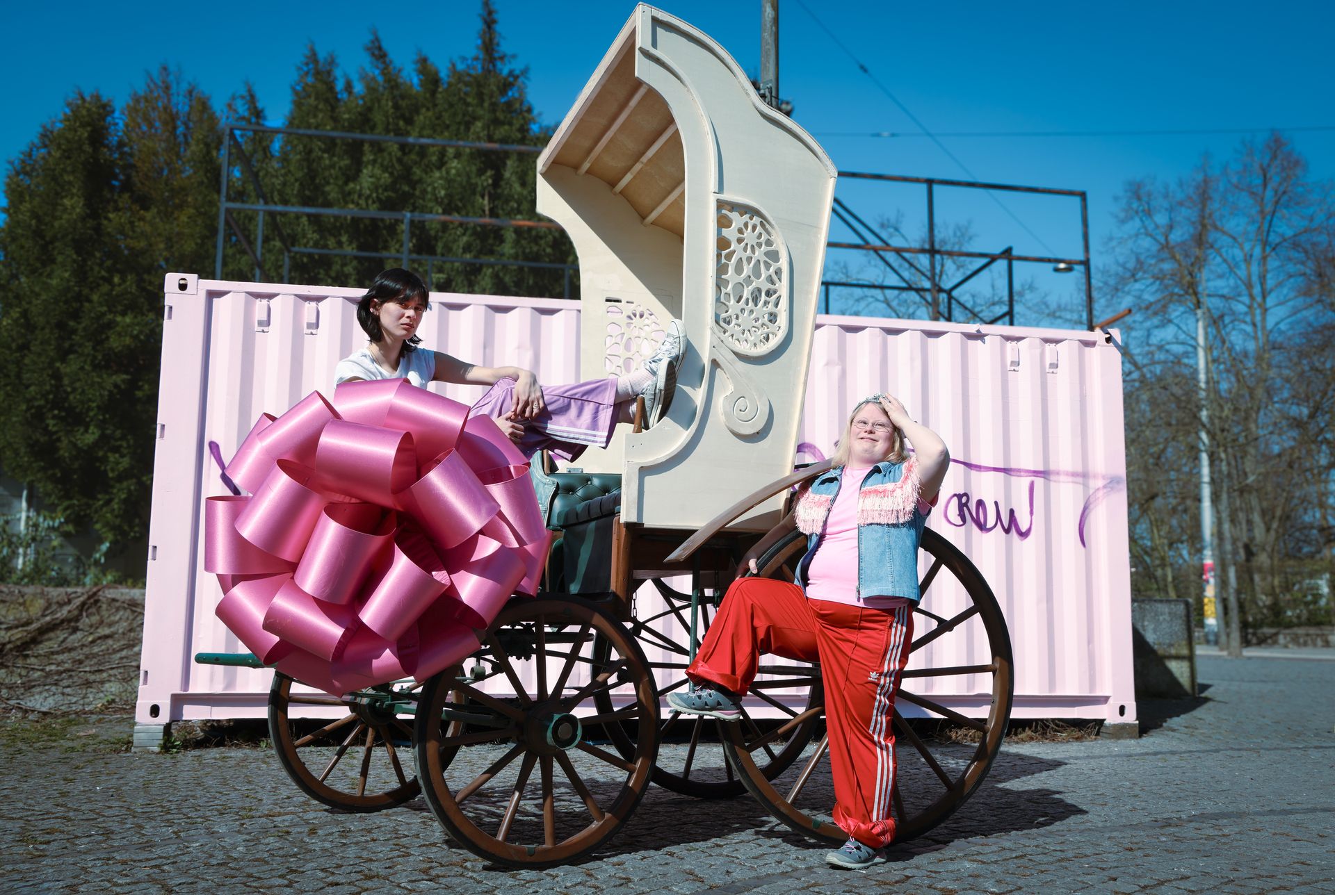 Im Freien steht ein rosa Container, davor eine Barock wirkende Kutsche mit Rädern aus dunklem Holz. Zwei Personen lehnen lässig an der Kutsche, links hängt an ihr eine gigantische pinke Satinschleife, wie sonst auf Geschenken zu finden.