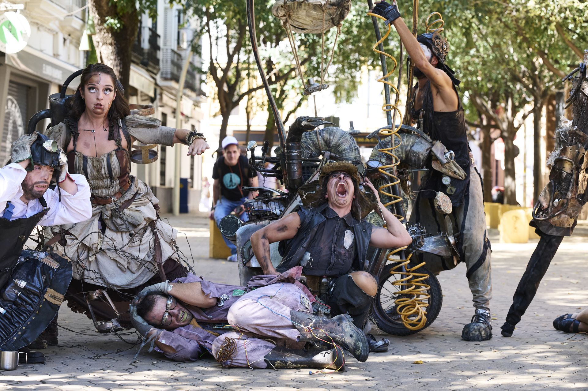 Sechs Personen sind auf dem Bild zu sehen, mehrere von ihnen schlagen die Hände über dem Kopf zusammen, sie wirken verzweifelt. Ihre Kostüme sind schmutzig, sie sehen nach Apokalypse und Steampunk aus.