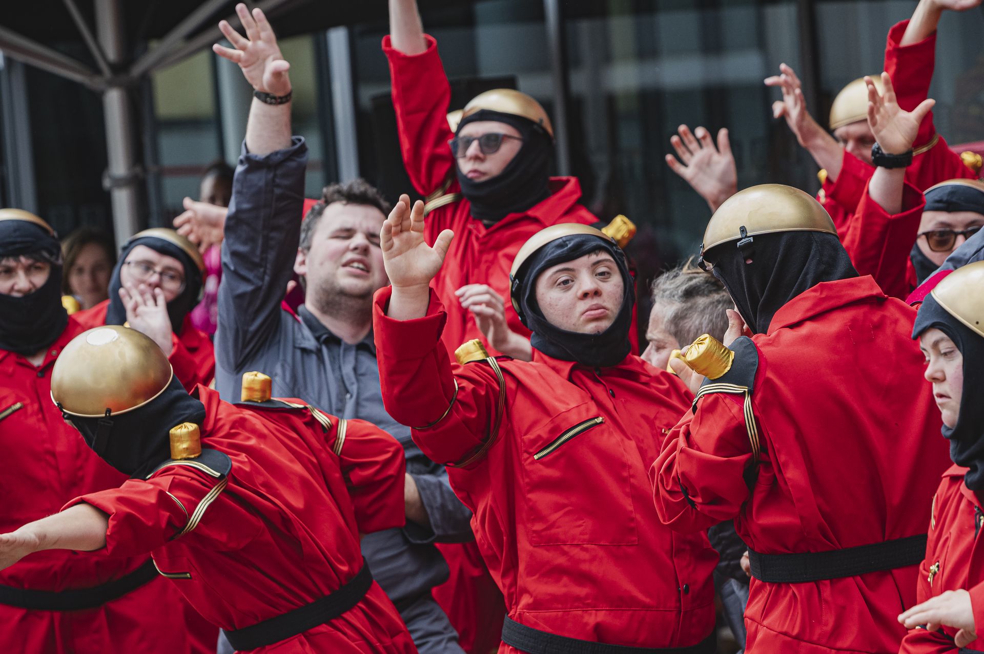 Elf Personen tanzen im Bild, sie stehen eng zusammen. Zwei von ihnen tragen graue Overalls, die übrigen rote Overalls und eine goldene Metallschale auf dem Kopf. Sie sind Roboter. Beim Tanzen strecken einigen die Arme vom Körper weg.